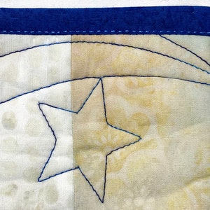May include: A close-up of a handmade quilt block with a blue star and a curved line stitched in blue thread. The quilt block is made of two different fabrics, one white and one beige.