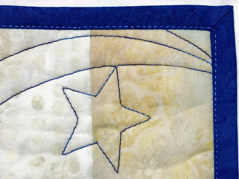 May include: A close-up of a handmade quilt block with a blue star and a curved line stitched in blue thread. The quilt block is made of two different fabrics, one white and one beige.