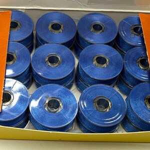 May include: A cardboard box containing twelve circular spools of blue sewing machine thread. The spools are arranged in a grid pattern, each featuring a clear plastic center core. The box has orange side flaps and a yellow base.