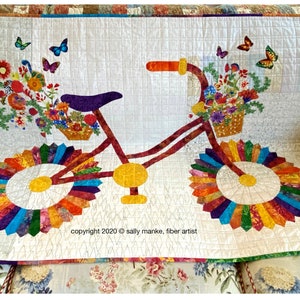Puede incluir: Una colcha colorida que representa una bicicleta con cestas llenas de flores y mariposas. Las ruedas de la bicicleta est&aacute;n hechas de piezas de tela de colores dispuestas en forma de sol. La colcha est&aacute; firmada "copyright 2020 &copy; sally manke, fiber artist".