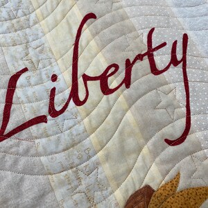 May include: A close-up of a beige fabric with a red cursive word "Liberty" stitched on it. The fabric has a pattern of small white stars and a quilted design.