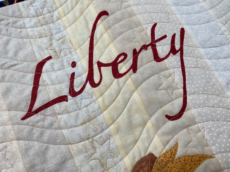 May include: A close-up of a beige fabric with a red cursive word "Liberty" stitched on it. The fabric has a pattern of small white stars and a quilted design.