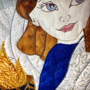 May include: A close-up of a quilted fabric artwork featuring a woman with brown hair and blue eyes. She is wearing a blue dress and has a golden flame behind her.