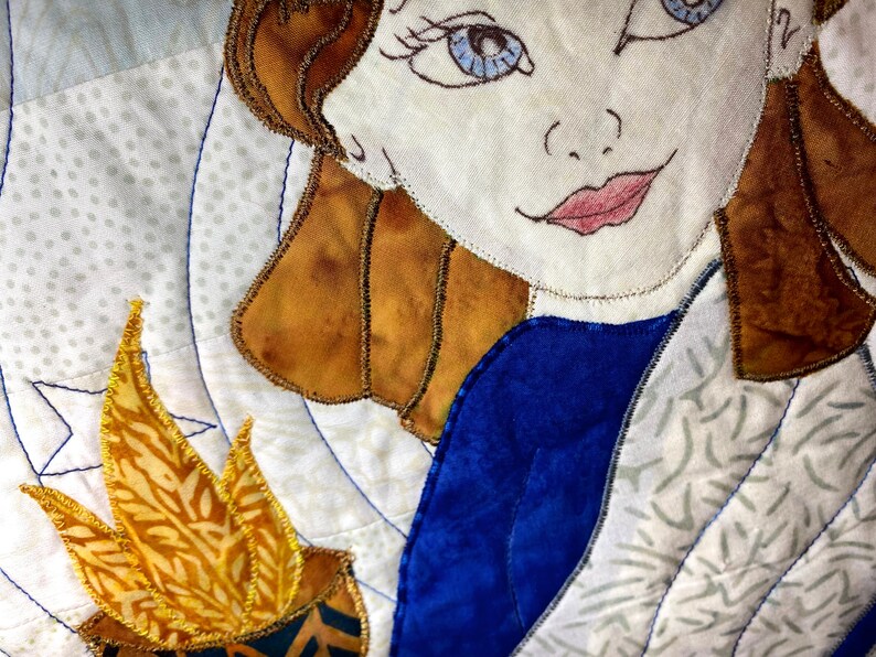 May include: A close-up of a quilted fabric artwork featuring a woman with brown hair and blue eyes. She is wearing a blue dress and has a golden flame behind her.