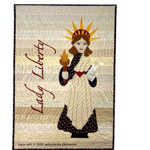 May include: A quilted wall hanging featuring a stylized image of Lady Liberty holding a torch and a document. The background is a gradient of beige and gray with a wavy pattern. The text "Lady Liberty" is written in red cursive.
