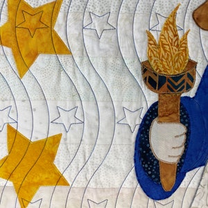 May include: A quilted fabric wall hanging with a blue, white, and gold color scheme. The design features a stylized figure holding a torch, surrounded by stars.
