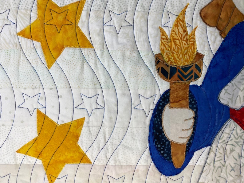 May include: A quilted fabric wall hanging with a blue, white, and gold color scheme. The design features a stylized figure holding a torch, surrounded by stars.