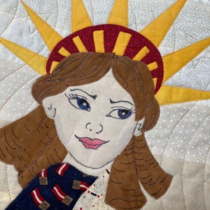 May include: A close-up of a quilted fabric artwork depicting a woman wearing a crown with a red and white striped band. The woman has brown hair and blue eyes. The background is a light beige fabric with a white star pattern.