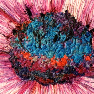 May include: Close-up of a colourful fabric flower with a textured centre. The centre is made of various shades of blue, red, and purple fabric pieces. The petals are a soft pink colour with a subtle shimmer.