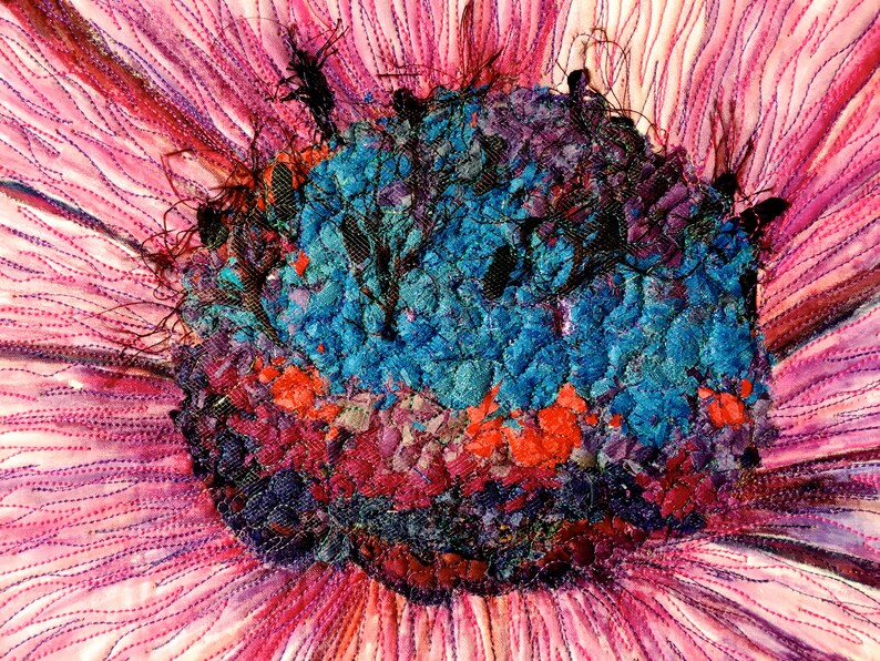 May include: Close-up of a colourful fabric flower with a textured centre. The centre is made of various shades of blue, red, and purple fabric pieces. The petals are a soft pink colour with a subtle shimmer.