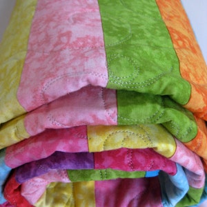 May include: A colorful striped quilt is rolled up, showcasing a variety of bright colors including pink, green, yellow, orange, and purple. The quilt is quilted with a decorative stitch pattern.