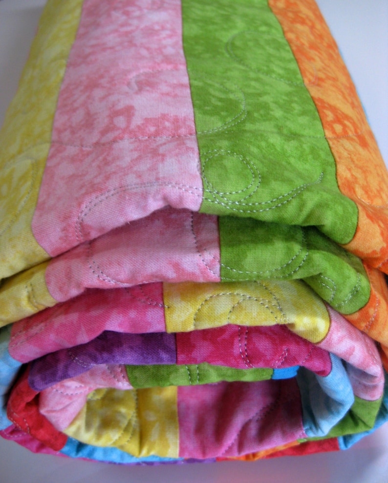 May include: A colorful striped quilt is rolled up, showcasing a variety of bright colors including pink, green, yellow, orange, and purple. The quilt is quilted with a decorative stitch pattern.