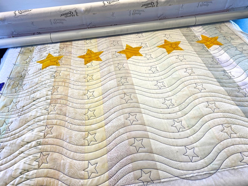 May include: A white quilt with a wavy pattern and five large yellow stars. The quilt is being quilted on a long arm quilting machine.