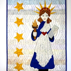 May include: A quilted wall hanging featuring a stylized depiction of the Statue of Liberty holding a torch and a book. The background is a white and blue wavy pattern with yellow stars.