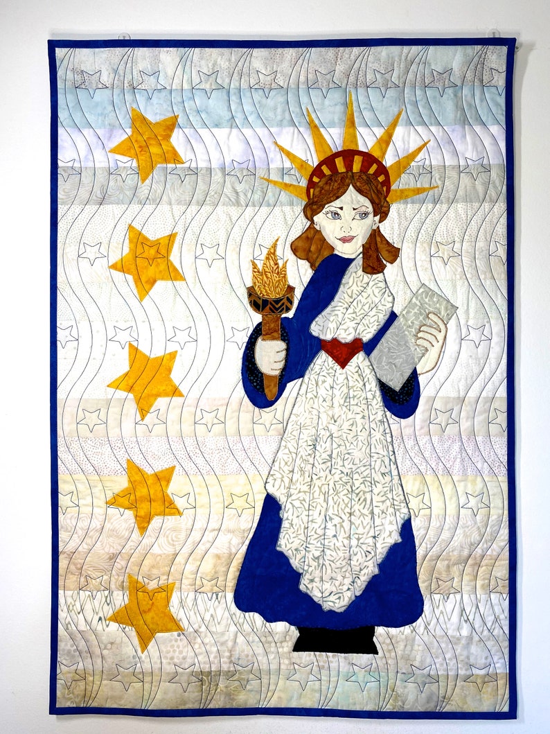 May include: A quilted wall hanging featuring a stylized depiction of the Statue of Liberty holding a torch and a book. The background is a white and blue wavy pattern with yellow stars.