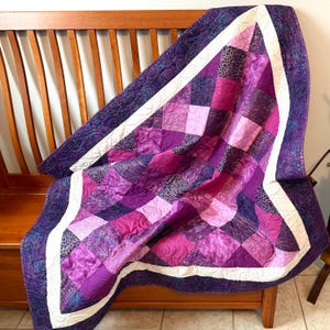 May include: A handmade quilt in shades of purple, magenta, and pink, with a white border. The quilt features a patchwork design with various fabric patterns and textures. The quilt is draped over a wooden bench.