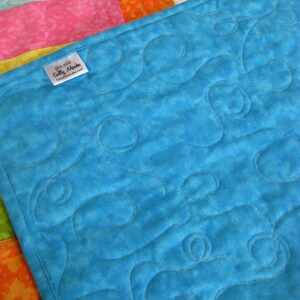 May include: A turquoise blue quilt with a swirling pattern quilted into the fabric. The quilt is backed with a white fabric with a floral pattern. A small white label with the text "Etsy shop Sally Makes" is attached to the quilt.
