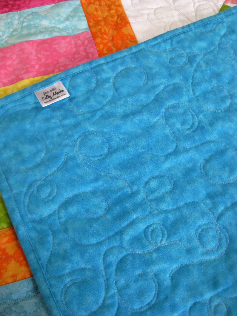 May include: A turquoise blue quilt with a swirling pattern quilted into the fabric. The quilt is backed with a white fabric with a floral pattern. A small white label with the text "Etsy shop Sally Makes" is attached to the quilt.