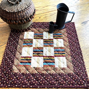 May include: A quilted table runner with a brown, beige, and blue patchwork design. The runner has a dark brown border with a small white floral pattern.