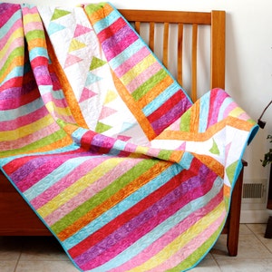 May include: A colorful striped quilt with a white background and a geometric pattern of triangles. The quilt is folded and draped over a wooden bench.