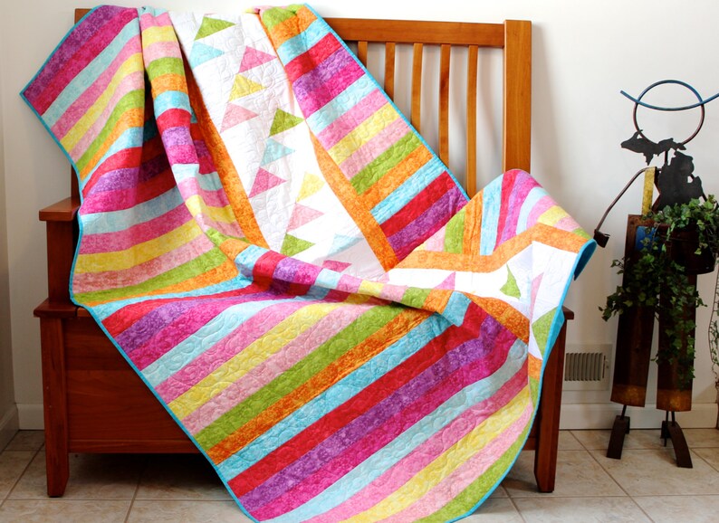 May include: A colorful striped quilt with a white background and a geometric pattern of triangles. The quilt is folded and draped over a wooden bench.