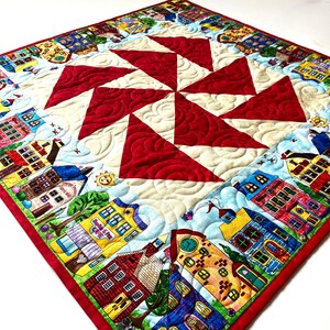 May include: A red and white quilt with a pinwheel design in the center. The border of the quilt features a colorful pattern of houses and buildings.