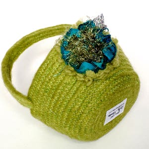 May include: A small green crocheted basket with a teal and gold fabric flower on top. The basket has a handle and a white label that says "Stitch Love".