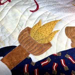 May include: A close-up of a quilted fabric design featuring a torch with a brown handle and a yellow flame. The torch is surrounded by a blue and red patterned fabric with stars.
