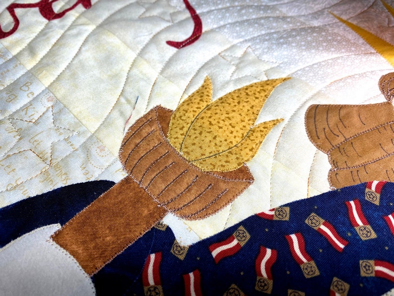 May include: A close-up of a quilted fabric design featuring a torch with a brown handle and a yellow flame. The torch is surrounded by a blue and red patterned fabric with stars.