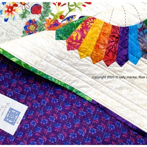 Puede incluir: Una colcha blanca con un dise&ntilde;o de abanico de colores arco&iacute;ris en el centro. La colcha est&aacute; respaldada con una tela morada con un patr&oacute;n floral. La colcha est&aacute; etiquetada con el texto "WIMSICAL Bicycle" y "Sally Manke fiber artist" y el a&ntilde;o de copyright 2020.