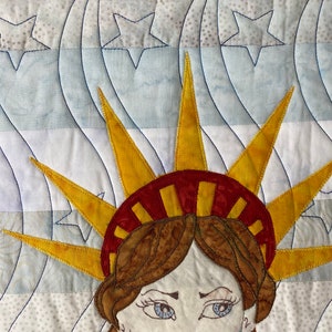 May include: A close-up of a quilted fabric depicting the Statue of Liberty. The fabric is white with blue and gray wavy lines and white stars. The Statue of Liberty is made of yellow, red, and brown fabric. Her face is visible with blue eyes and brown hair.