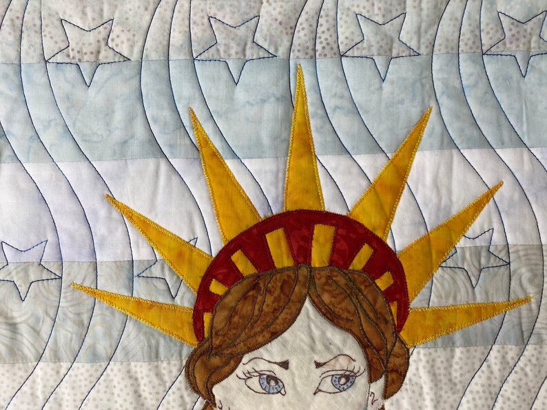 May include: A close-up of a quilted fabric depicting the Statue of Liberty. The fabric is white with blue and gray wavy lines and white stars. The Statue of Liberty is made of yellow, red, and brown fabric. Her face is visible with blue eyes and brown hair.