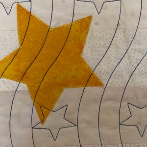 May include: A yellow star with a stitched border is sewn onto a white fabric background with a pattern of small white dots. The star is layered over a design of curved lines stitched in black thread.