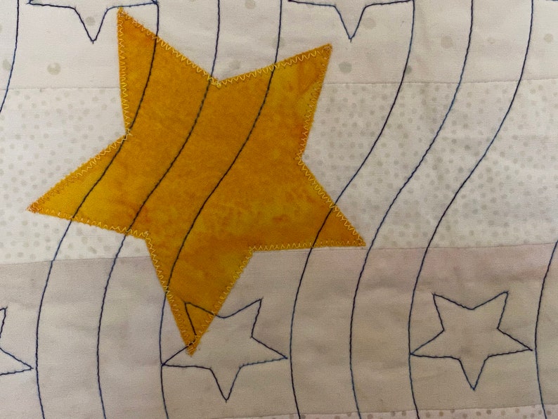 May include: A yellow star with a stitched border is sewn onto a white fabric background with a pattern of small white dots. The star is layered over a design of curved lines stitched in black thread.