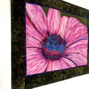 May include: A close-up of a pink flower with a purple and blue centre. The flower is framed in a dark green and brown fabric.