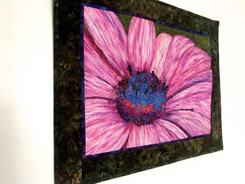 May include: A close-up of a pink flower with a purple and blue centre. The flower is framed in a dark green and brown fabric.