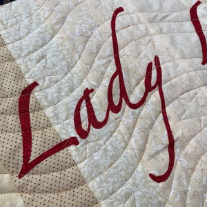 May include: A close-up of a beige and brown quilted fabric with the word "Lady" embroidered in red cursive lettering.