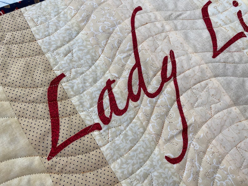 May include: A close-up of a beige and brown quilted fabric with the word "Lady" embroidered in red cursive lettering.