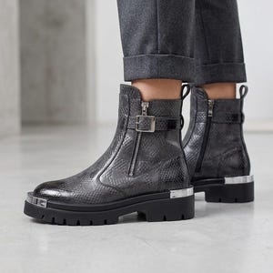 Könnte beinhalten: Dunkelgraue Stiefeletten mit Schlangenmuster. Die Stiefel haben einen Reißverschluss, eine Schnalle und eine dicke schwarze Sohle mit silbernen Akzenten. Getragen mit grauer Hose.