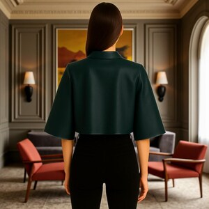 Pu&ograve; includere: Giacca corta verde scuro con maniche ampie e colletto. La giacca &egrave; abbinata a pantaloni neri. L'immagine &egrave; scattata in interni, con un dipinto e lampade visibili sullo sfondo.