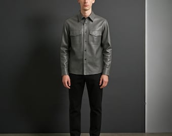 May include: A gray leather shirt with two chest pockets and a button-down front. The shirt is paired with black pants and black shoes. The model is standing in front of a dark gray background.