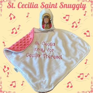 May include: A white and pink plush security blanket featuring a soft, textured minky fabric on the underside. The top centre displays an embroidered figure of St. Cecilia with a halo, and the text reads: St. Cecilia pray for Cecilia Therese!