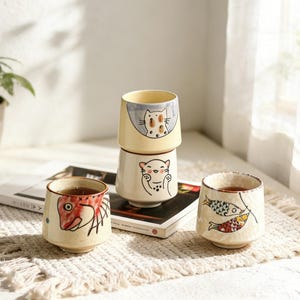 Könnte beinhalten: Ein Set aus drei Keramikbechern mit gesprenkelter, matter Oberfläche. Die Becher sind in Creme-, Hellgrau- und Dunkelgrautönen gehalten. Jeder Becher hat ein schlichtes, minimalistisches Design, ideal für Kaffee oder Tee.