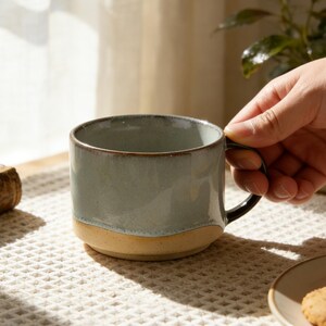 May include: A ceramic mug with a two-tone design. The upper portion is a muted blue-green, while the bottom is a tan color. The mug has a black handle and is held by a hand. The mug is on a white waffle-weave surface.