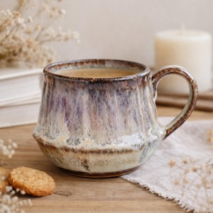 Könnte beinhalten: Eine handgefertigte Keramik-Tasse mit einer einzigartigen Glasur in Braun-, Lila- und Cremetönen. Die Tasse ist mit einer hellbraunen Flüssigkeit gefüllt, wahrscheinlich Kaffee oder Tee. Sie steht auf einer Holzoberfläche mit Keksen und einer Kerze im Hintergrund.