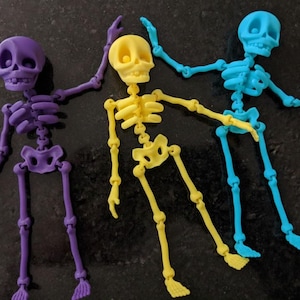 May include: Three articulated skeleton figures in purple, yellow, and blue. Each skeleton has a skull, rib cage, and articulated limbs. The skeletons are posed in a playful manner, with arms raised.