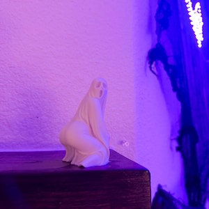 May include: A small, off-white ghost figurine with a simple, stylized design. The ghost is kneeling on a dark wooden surface, with a textured white wall in the background. The scene is lit with purple and blue hues, creating a spooky atmosphere.
