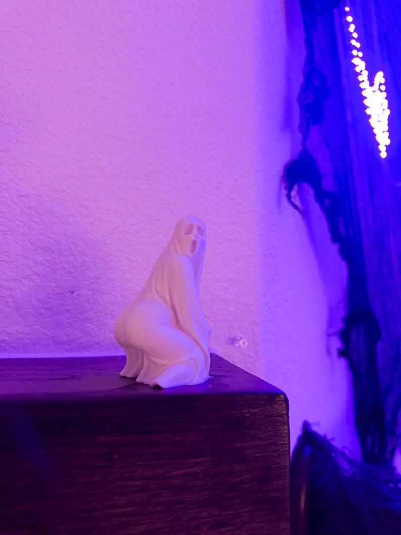 May include: A small, off-white ghost figurine with a simple, stylized design. The ghost is kneeling on a dark wooden surface, with a textured white wall in the background. The scene is lit with purple and blue hues, creating a spooky atmosphere.