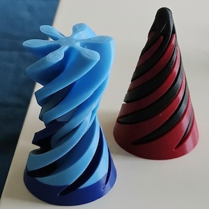 May include: Two abstract 3D-printed sculptures. One is a blue, spiraling form with a star-like top. The other is a red and black striped cone shape. Both are on a white surface.