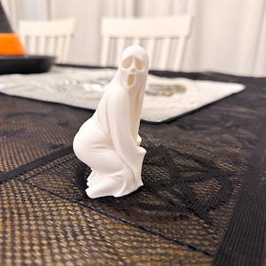 May include: A white ghost figurine in a kneeling pose, with a wide open mouth and eye sockets. The ghost is on a black mesh surface, with a table setting in the background. The figurine is a Halloween decoration.